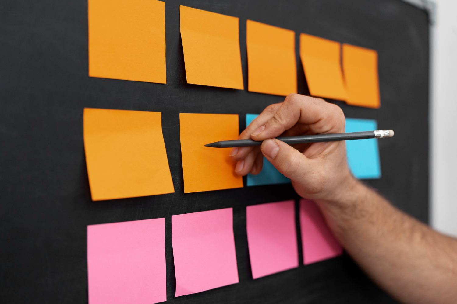 A hand holding a pencil points to colorful sticky notes on a blackboard, symbolizing task organization—visualizing how to delegate as a leader effectively.