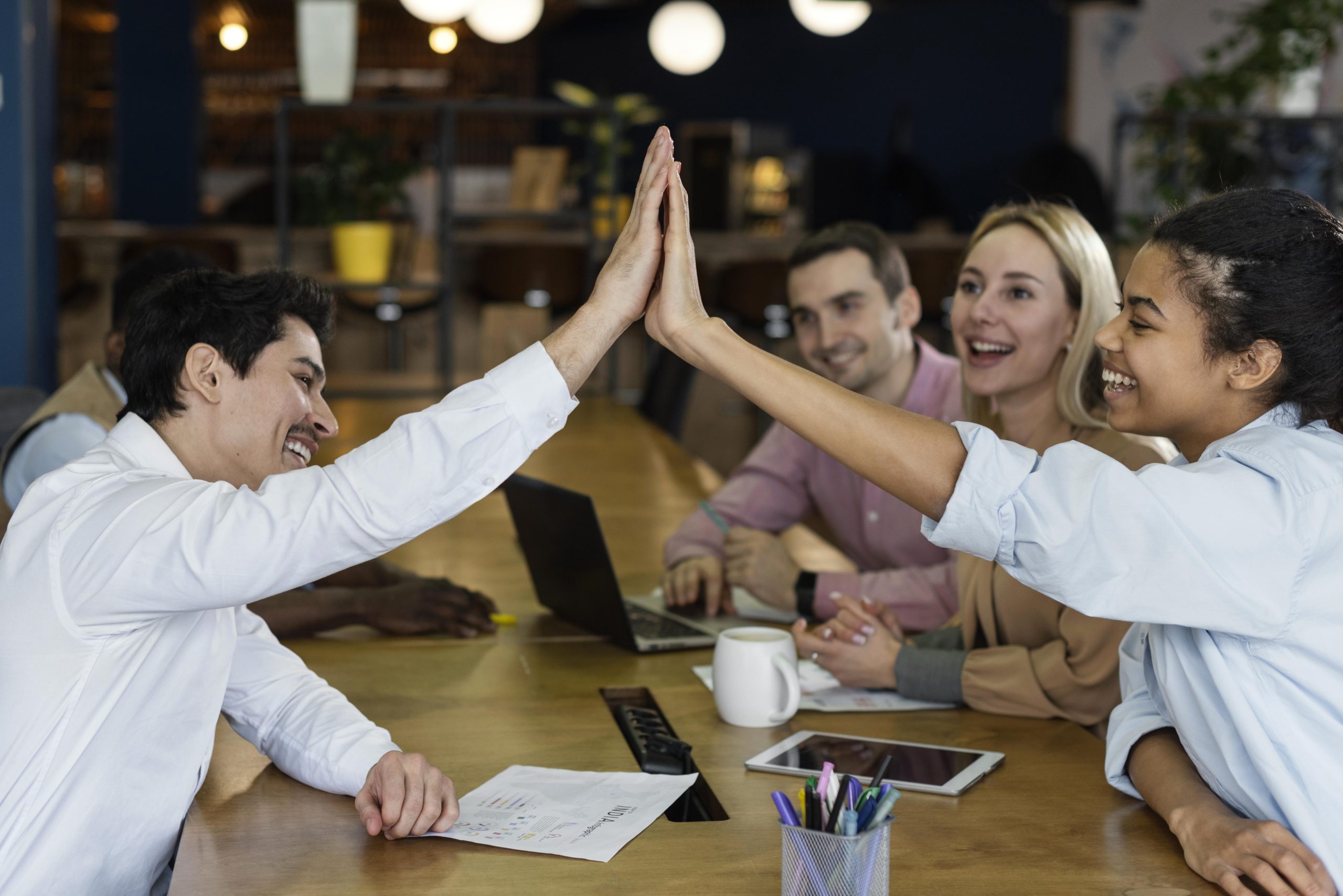 A group of co-workers having a good time around a table, with two of them highfiving each other, symbolizing team work and communication. This explains the necessity of tracking and improving employee engagement metrics with training.
