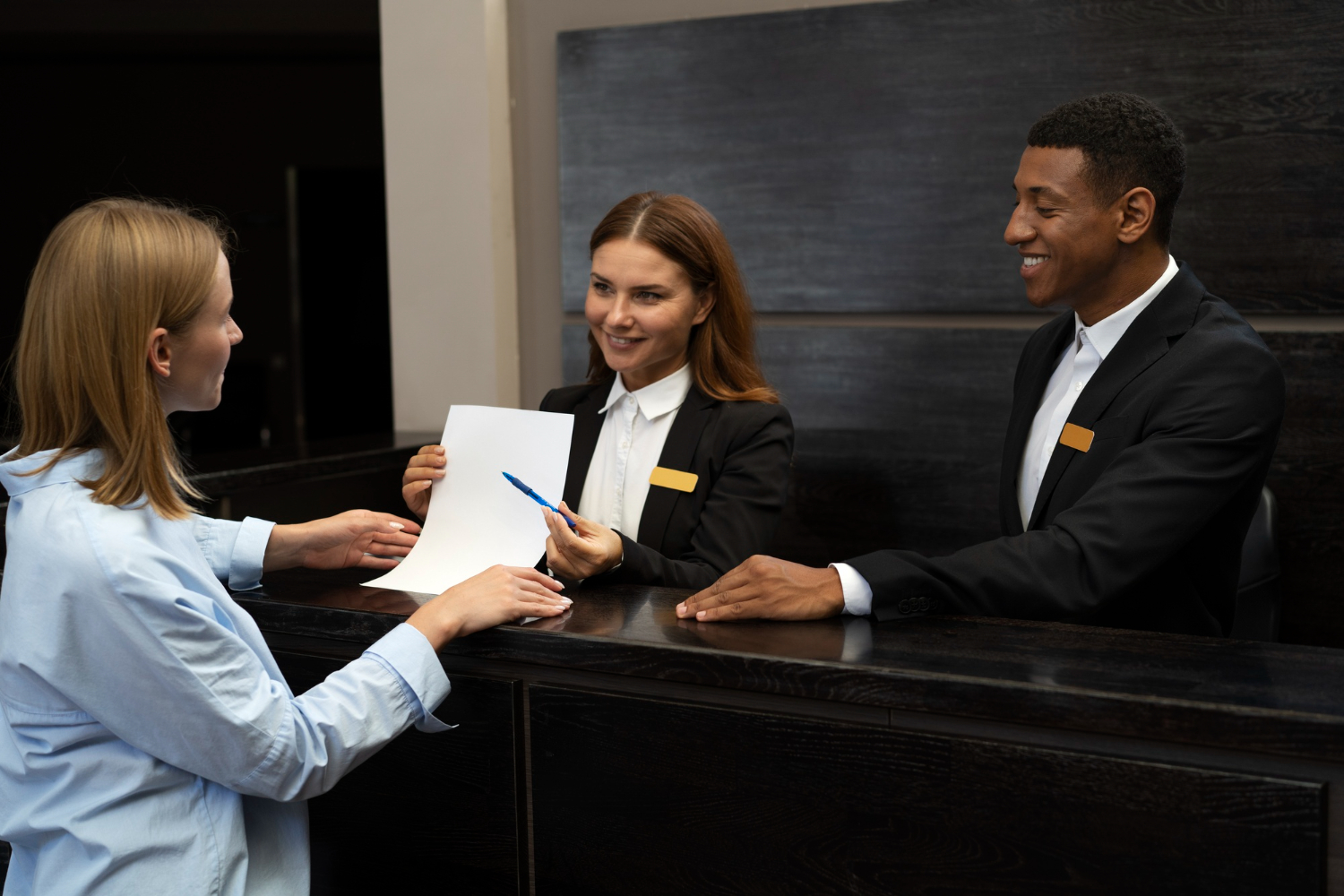 Two receptionists talking to a customer at a hotel’s front desk, smiling and offering them to sign a document, which shows the importance of providing training for reception staff.