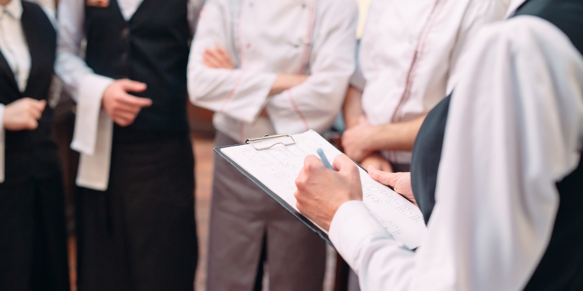 Training restaurant staff in a pre-shift meeting, with a manager holding a clipboard and giving instructions to chefs and waitstaff.
