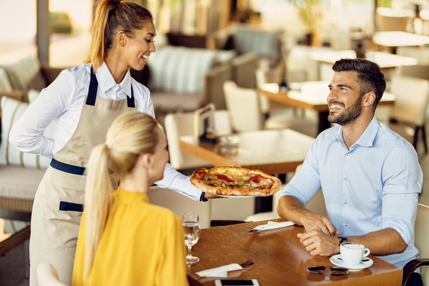 A smiling waitress serves a fresh pizza to cheerful guests in a modern restaurant, reflecting the results of effective hospitality customer service training.