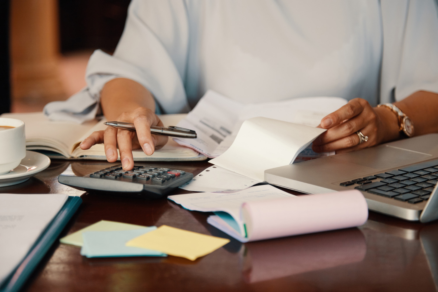 Person calculating expenses with a calculator, notebook, and laptop while reviewing documents, representing planning a learning and development budget.